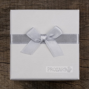 Puede incluir: Una caja de regalo blanca con una cinta y un lazo plateados. La caja tiene el texto "PROZART" impreso en la parte delantera.