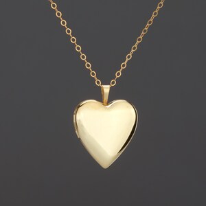 May include: A polished gold heart-shaped locket pendant suspended from a delicate gold chain. The locket features a smooth, reflective surface with a classic heart silhouette and a small hinge on the left side, showcasing a clean, minimalist design.