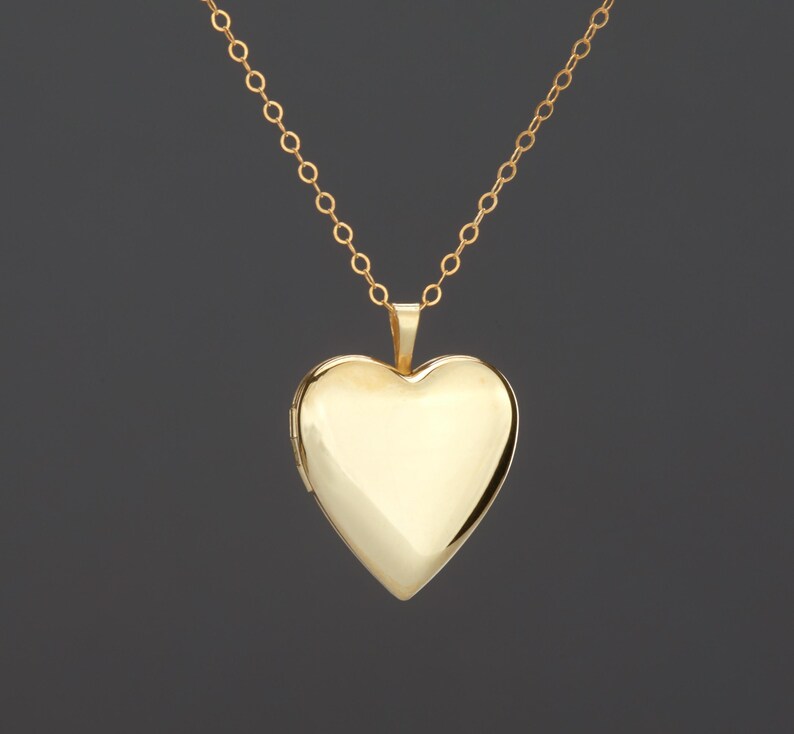 May include: A polished gold heart-shaped locket pendant suspended from a delicate gold chain. The locket features a smooth, reflective surface with a classic heart silhouette and a small hinge on the left side, showcasing a clean, minimalist design.