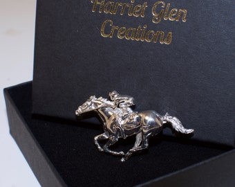 Puede incluir: Figurita de plata de un caballo y un jinete, una escultura detallada de un caballo en carrera con un jinete montado en su lomo. La figurita se muestra en una caja negra con el texto "Harriet Glen Creations" en la tapa.