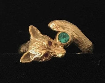 May include: A gold ring featuring a detailed fox design with a green gemstone set in the center. The fox's eyes are accented with red gemstones.