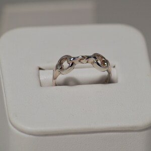 Könnte beinhalten: Ein silberner Ring mit einem Kettenglied-Design. Der Ring besteht aus einem glänzenden, metallischen Material und weist eine Reihe von miteinander verbundenen Gliedern auf, die ein durchgehendes Band bilden. Der Ring wird auf einer weißen Oberfläche präsentiert.