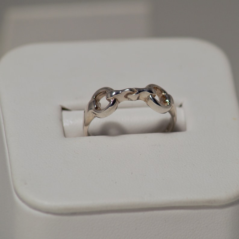 Puede incluir: Un anillo plateado con un dise&ntilde;o de eslabones de cadena. El anillo est&aacute; hecho de un material met&aacute;lico brillante y presenta una serie de eslabones interconectados que forman una banda continua. El anillo se muestra sobre una superficie blanca.