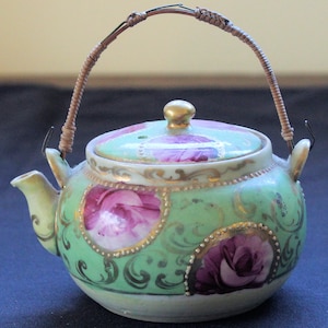 May include: A small, green and white porcelain teapot with a floral design and a wire handle. The teapot has a lid with a small hole in the top.