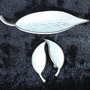 Puede incluir: Un conjunto de broche y pendientes plateados con forma de hojas. El broche es una hoja larga y delgada con vetas detalladas, y los pendientes son hojas curvas más pequeñas. El conjunto se muestra sobre una superficie oscura y texturizada.