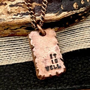 May include: A copper-colored pendant necklace with a rectangular charm. The charm has the words "IT IS WELL" stamped on it. The necklace chain is also copper-colored and has a link design. The background includes a textured surface.