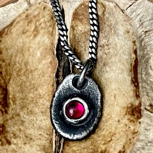 May include: A silver pendant necklace with a textured, oval-shaped pendant. The pendant features a bezel-set, round, red gemstone. The necklace chain is a silver, woven design. The jewelry is displayed on a textured, light brown surface.