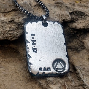 May include: A silver-colored rectangular pendant with a textured surface, hanging from a black chain. The pendant has the date "8.14.17" and the letters "RAM" engraved on it, along with a triangle symbol.