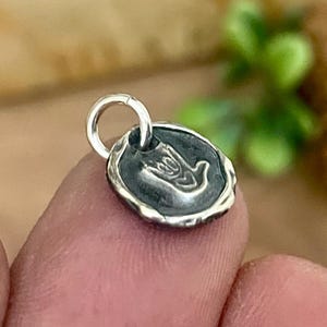May include: A small, round silver charm with a raised design of a hand making the "I love you" sign. The charm has a textured border and a small loop for attaching to a necklace or bracelet. The charm is held between two fingers.