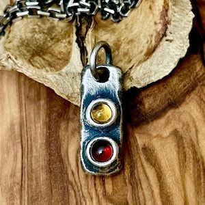 May include: A silver pendant necklace featuring a rectangular pendant with two round, colorful gemstones. The top gemstone is yellow, and the bottom is red. The pendant hangs from a silver chain.