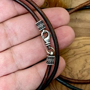 May include: Close-up of a brown leather necklace with a silver clasp and decorative silver beads. The necklace has two strands of leather and a lobster claw clasp. The silver beads have a repeating wave pattern.