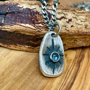 May include: A silver-toned pendant necklace with a textured, oval-shaped pendant. The pendant features a starburst design with a central blue gemstone. The necklace has a silver chain.