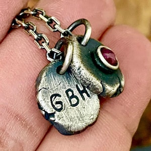 May include: A silver pendant necklace featuring two charms. One charm is a textured oval with the initials "GBH" engraved. The other charm is a small, textured oval with a red gemstone. The necklace chain is also silver.