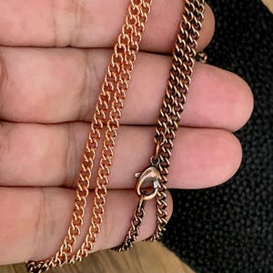 May include: Close-up of two rose gold chain necklaces. One chain is a lighter shade of rose gold, while the other is a darker, more bronze-like color. The necklaces are linked together with a lobster clasp.