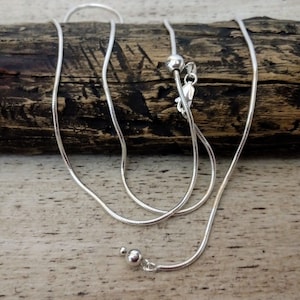 May include: A silver snake chain necklace with a sliding clasp. The chain is laying on a piece of wood.