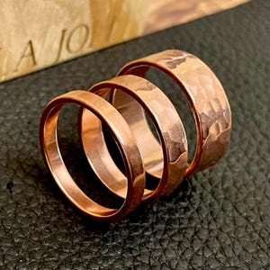 May include: Three copper rings of varying widths are displayed. The rings have a warm, reddish-brown hue. The widest ring has a hammered texture, while the others are smooth. The rings are arranged on a dark, textured surface.