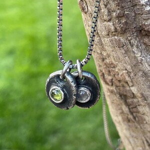 May include: A silver necklace featuring two small, textured pendants. Each pendant has a bezel-set gemstone: one green and one clear. The necklace chain is a box link style, and the background is a blurred green.