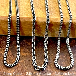 May include: Three silver chain necklaces with different link styles: a 2mm curb chain, a 2.5mm anchor chain, and a 1.6mm box chain. The chains are displayed on a brown wooden surface. The text "2mm Curb", "2.5mm Anchor", and "1.6mm box" is visible.