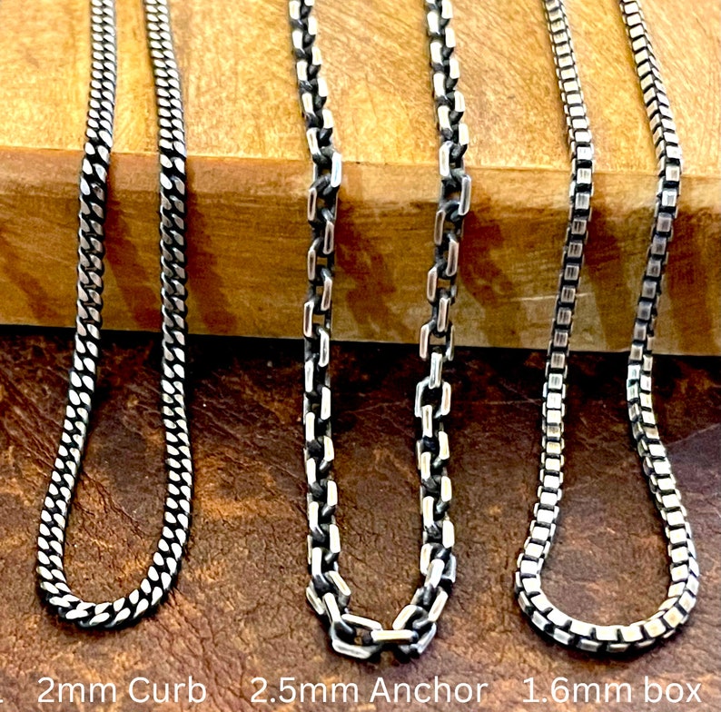 May include: Three silver chain necklaces with different link styles: a 2mm curb chain, a 2.5mm anchor chain, and a 1.6mm box chain. The chains are displayed on a brown wooden surface. The text "2mm Curb", "2.5mm Anchor", and "1.6mm box" is visible.
