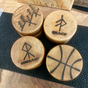 May include: Four wooden coasters with unique designs. One coaster features a tree design, another has a symbol with the word "GRACE", a third has a symbol with "COURAGE", and the last has a basketball graphic. The coasters are made of light brown wood.