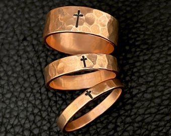 Puede incluir: Tres anillos de color cobre con una textura martillada y un diseño de cruz negra. Los anillos están apilados sobre una superficie negra, mostrando sus diferentes anchos y la detallada artesanía.