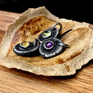 May include: Three handcrafted silver pendants with gemstone accents. Each pendant features a unique gemstone: yellow, green, and purple. The pendants are displayed on a textured, brown surface, showcasing the jewelry's rustic charm and design.