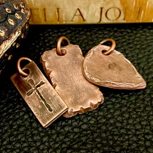 May include: Three copper pendants with a rustic finish. One pendant is rectangular with a cross design, another is a textured rectangle, and the third is leaf-shaped. Each has a loop for a chain.