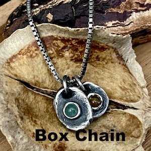 May include: A silver box chain necklace with two pendant charms. One charm features a green stone, and the other has a yellow stone. The necklace is displayed on a textured, light brown surface. The text "Box Chain" is at the bottom.
