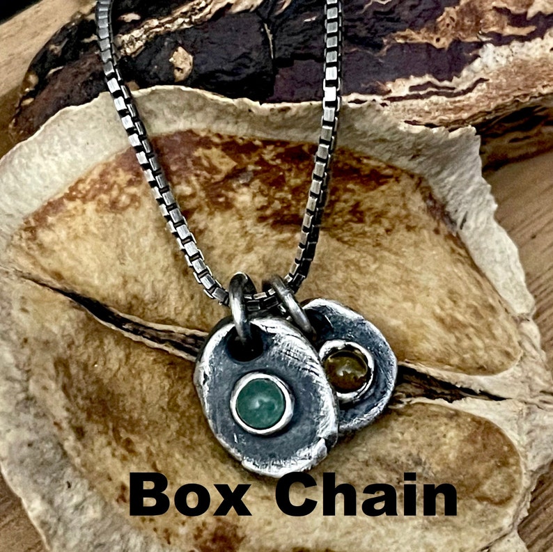 May include: A silver box chain necklace with two pendant charms. One charm features a green stone, and the other has a yellow stone. The necklace is displayed on a textured, light brown surface. The text "Box Chain" is at the bottom.
