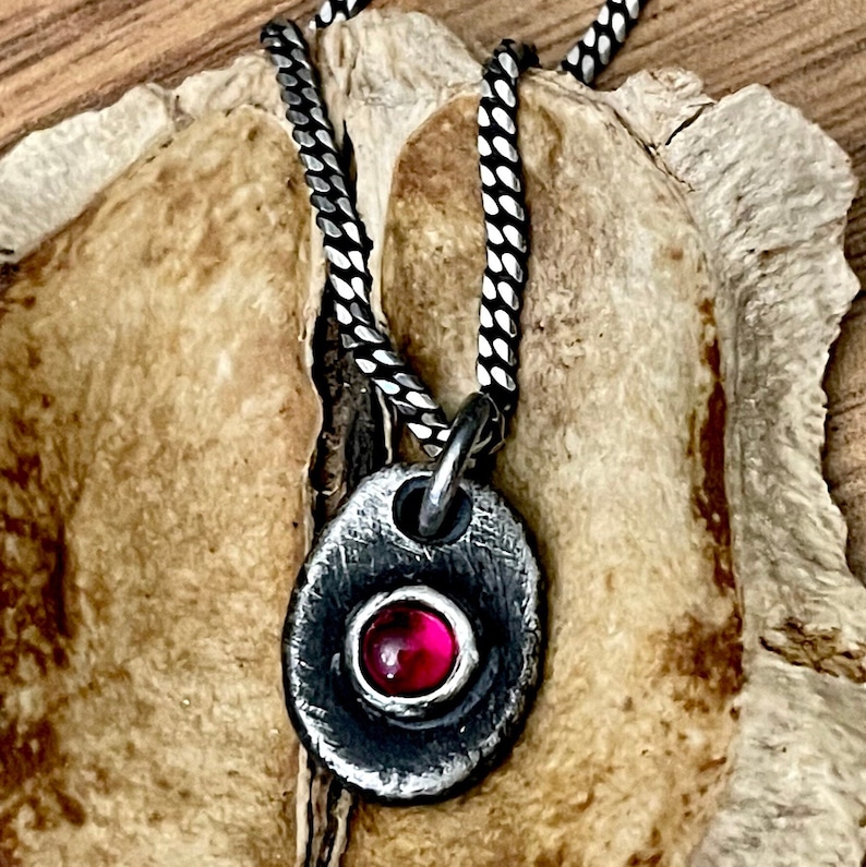 May include: A silver pendant necklace with a textured, oval-shaped pendant. The pendant features a bezel-set, round, red gemstone. The necklace chain is a silver, woven design. The jewelry is displayed on a textured, light brown surface.