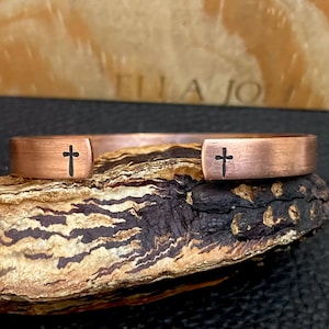 May include: A copper cuff bracelet with a cross etched on each end. The bracelet has a brushed finish and is displayed on a textured, multi-toned surface. The background features the text "ELLA JO".