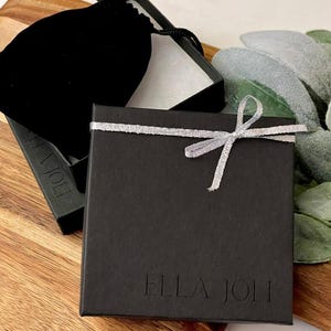 May include: A black gift box with a silver ribbon tied in a bow. The box is on a wooden surface, next to a black velvet pouch and a sprig of greenery. The text "ELLA JOLI" is printed on the box.