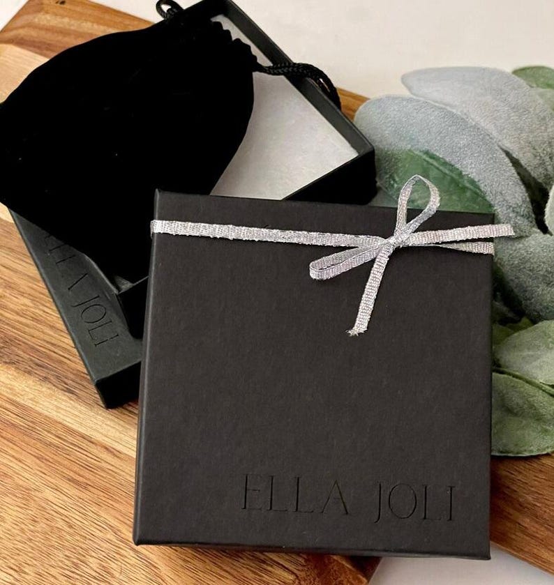 May include: A black gift box with a silver ribbon tied in a bow. The box is on a wooden surface, next to a black velvet pouch and a sprig of greenery. The text "ELLA JOLI" is printed on the box.