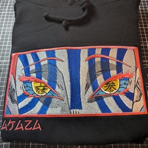 May include: Black hooded sweatshirt featuring an embroidered design of stylized eyes. The eyes have blue and white striped backgrounds, red outlines, and yellow and orange details. Japanese text is below the eyes.