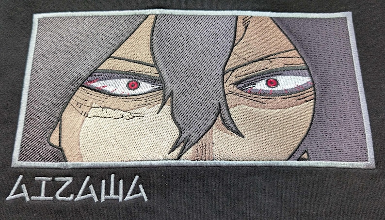 Puede incluir: Primer plano de una imagen bordada en blanco y negro de la cara de un personaje con ojos rojos. La imagen est&aacute; enmarcada en un borde azul claro. El texto "AIZAWA" est&aacute; bordado debajo de la imagen.