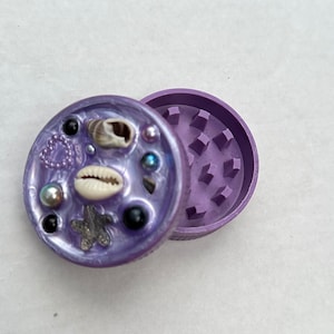Puede incluir: Un molinillo morado de dos piezas con una tapa decorativa. La tapa está adornada con conchas marinas, perlas y cuentas negras. El interior del molinillo tiene una superficie texturizada para moler.