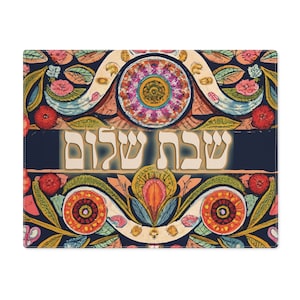 May include: A rectangular placemat with a floral design in shades of blue, pink, orange, and yellow. The words "Shabbat Shalom" are displayed in a cream-colored font across a dark blue banner. The placemat has a decorative, artistic style.