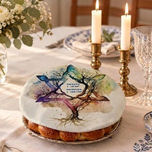 May include: A round, off-white fabric cover featuring a watercolor-style Star of David intertwined with a tree, with Hebrew text in the center. The cover sits atop a round tray filled with golden-brown, round bread rolls on a dining table.