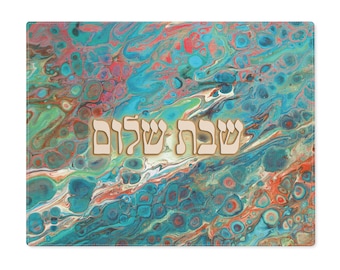 May include: A turquoise, green, and orange abstract patterned fabric placemat with the Hebrew words "Shabbat Shalom" in gold lettering.