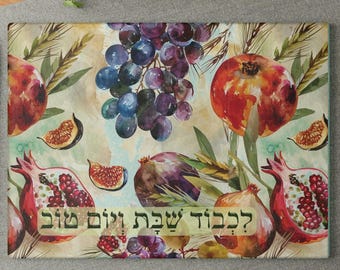 May include: Rectangular cutting board with a watercolor design featuring pomegranates, grapes, and figs. The fruits are in shades of red, purple, and brown, with green leaves and wheat stalks. The text in the image is in Hebrew.