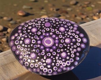 Una piedra de meditación mandala pintada a mano inspiradora con tonos púrpuras sobre un fondo negro profundo.