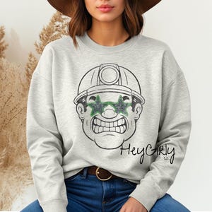 May include: Pale grey sweatshirt with a cartoon miner wearing star-shaped green sunglasses and a hard hat. The miner has a stern expression and a wide grin. The text "Hey Girly co." is written in cursive on the lower right.