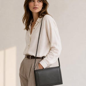 May include: A rectangular black crossbody bag with a braided edge detail and a thin shoulder strap. The bag features a structured flap closure and a smooth finish, worn across the body with a light-coloured button-down shirt and tan trousers.