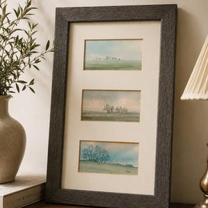 May include: A rectangular dark gray textured frame containing three separate landscape watercolor paintings stacked vertically. Each painting depicts a soft, hazy outdoor scene with trees, fields, and a pale sky, all set against a cream-colored mat board.