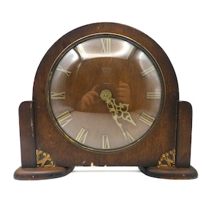 May include: A vintage wooden clock with a rounded top and a circular clock face. The clock face features Roman numerals and gold-colored hands. Decorative gold accents are on the base. The clock is a classic piece of home decor.