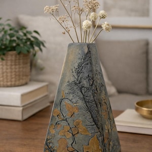 May include: A decorative, triangular vase with a blue-grey finish, featuring gold and black botanical designs. The vase holds dried, white flowers. The vase is on a wooden table.