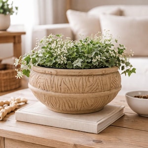 May include: A beige, textured ceramic planter filled with green and white leafy plants and small white flowers. The planter sits on a closed, light-coloured book on a wooden table. A white bowl is visible in the background.