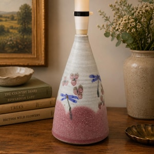 Könnte beinhalten: Ein konischer Keramik-Tischlampenfuß mit einem gesprenkelten cremeweißen oberen Teil und einem strukturierten altrosa Sockel. Die Oberfläche ist mit handgemalten blauen Libellen und kleinen rosa Blumenclustern verziert.