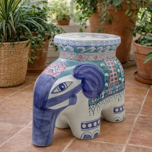 May include: A ceramic garden stool shaped like an elephant, featuring a white base with hand-painted blue, green, and pink floral and geometric patterns. The stool has a flat, circular top and a sturdy four-legged base with decorative motifs.
