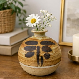 May include: A round, tan ceramic vase with a narrow neck and a flared rim. The vase features a dark brown, abstract leaf-like pattern painted on the upper half, separated by a thin white horizontal band from the speckled, light tan lower base.